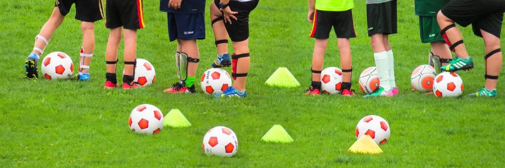 Soccer players stood on a training field with balls and cones on the ground. 