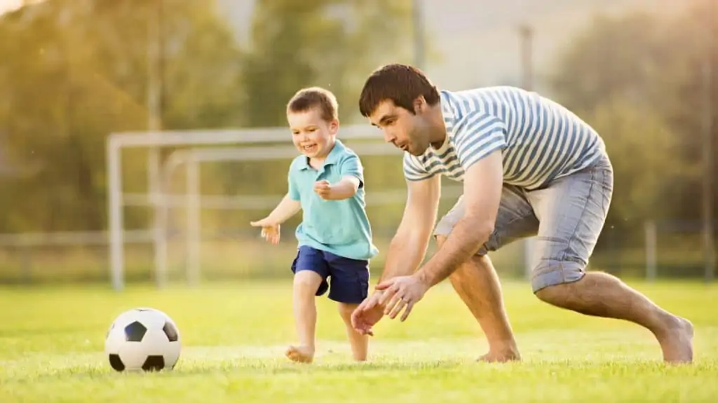 A 3-4-year-old kid playing soccer with dad, running after a ball